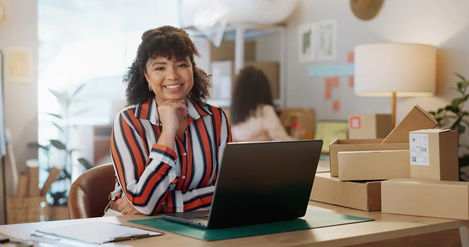 Vendedora sorridente no notebook, com caixas de pedidos ao fundo
